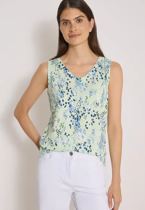 Frau trägt ein ärmelloses, hellgrünes Top mit blauem Blumenmuster und V-Ausschnitt zu weißen Hosen.