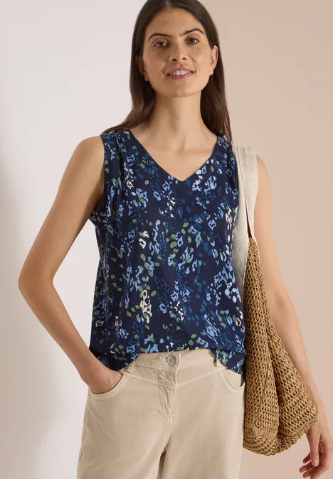Frau trägt blaues, ärmelloses Blumen-Top, beige Hose und geflochtene Tasche.
