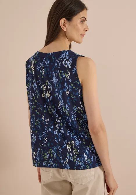 Frau von hinten im ärmellosen, dunkelblauen, floralen Top und beiger Hose vor hellem Hintergrund.
