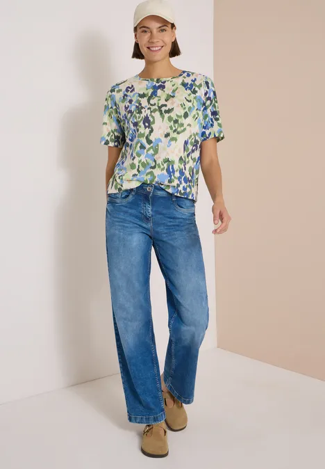 T-Shirt mit Boat Neck und Print cashmere beige Dame trägt eine gemusterte Bluse mit Knopfleiste, weite Jeans und beige Schuhe, lächelnd vor neutralem Hintergrund.