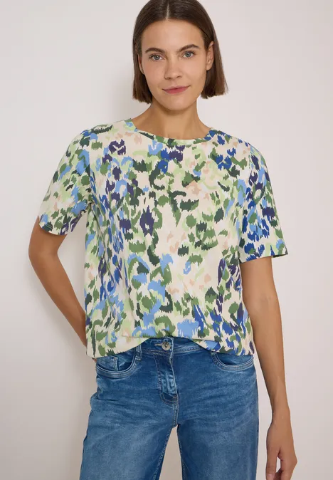 T-Shirt mit Boat Neck und Print cashmere beige Frau trägt bunt gemustertes Kurzarmshirt mit grün-blauem abstraktem Blumenprint und Jeans.