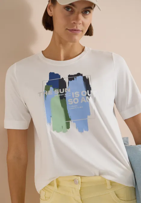Weißes T-Shirt mit blau-grünen Pinselstrichen und 'THE SUN IS OUT SO AM I'-Aufdruck, kombiniert mit gelber Hose.