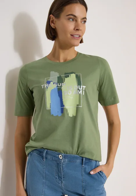 T-Shirt mit Rundhals und Frontprint deep matcha green Frau trägt grünes T-Shirt mit abstraktem Print und blaue Jeans, steht vor heller Wand mit Hand in Hosentasche.