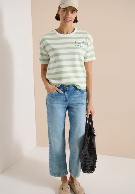 Vrouw in groen-wit gestreept shirt, blauwe culotte-jeans, beige pet en zwarte tas voor een neutrale achtergrond.