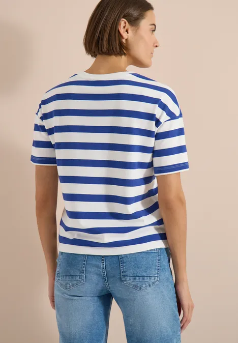 Achteraanzicht van een persoon in een blauw-wit gestreept t-shirt en spijkerbroek voor een beige muur.