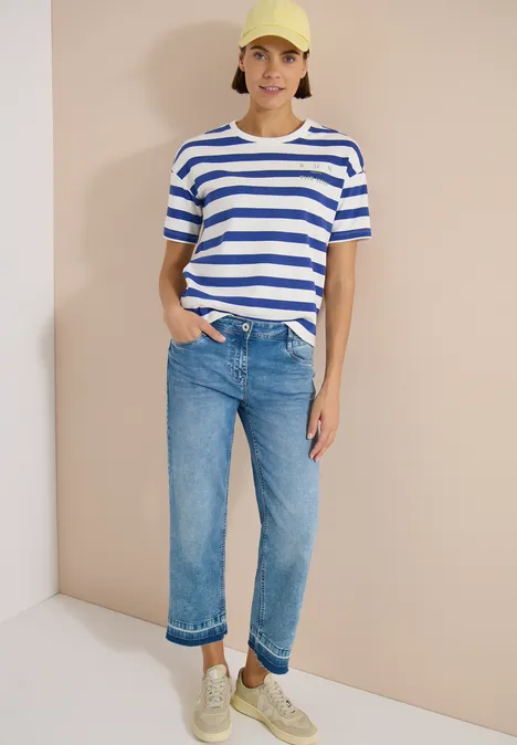 Vrouw met blauw-wit gestreept t-shirt, blauwe opgerolde jeans en witte sneakers tegen een neutrale achtergrond.