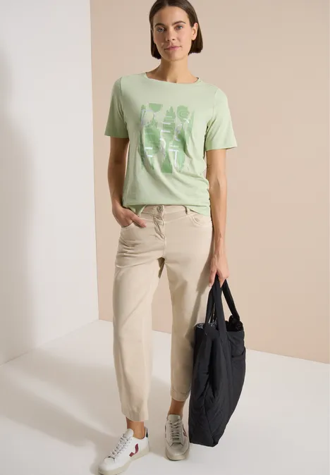 Frau trägt grünes T-Shirt mit Motiv, helle Hose, weiße Sneaker und dunkle Tasche vor neutralem Hintergrund.