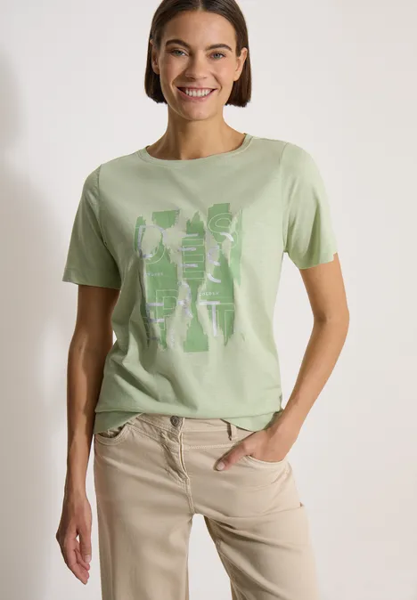 Frau trägt hellgrünes T-Shirt mit Grafikprint und beiger Hose, steht seitlich mit Hand in Tasche.