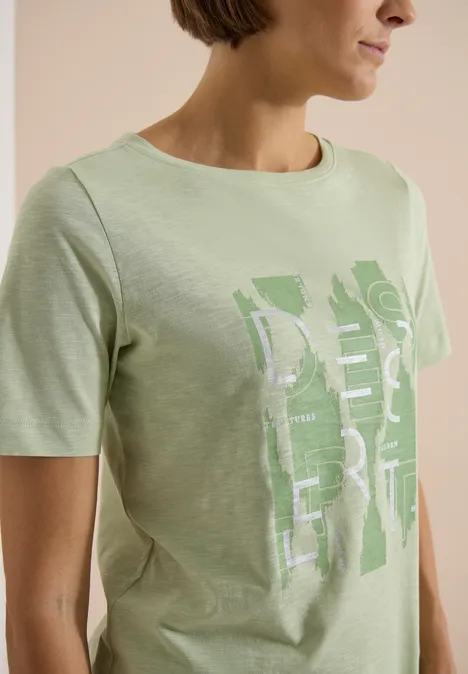 Frau in hellgrünem T-Shirt mit grafischem weißen/hellgrünen Print, vor schlichter beiger Hintergrund.