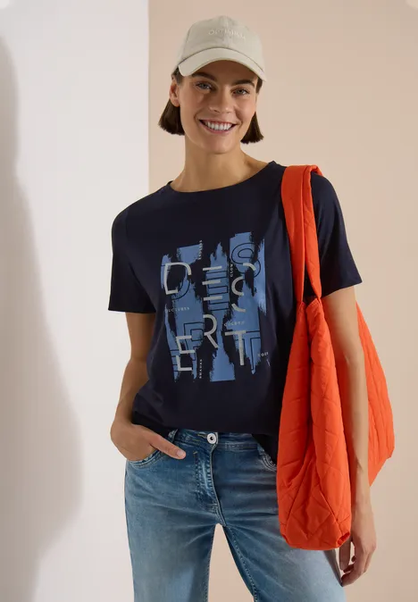 Person trägt Jeans, dunkelblaues Shirt mit Print, orange Umhängetasche und hellen Bucket Hat vor hellem Hintergrund.
