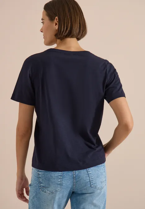 Person von hinten in dunkelblauem T-Shirt und hellblauen Jeans vor beigem Hintergrund.