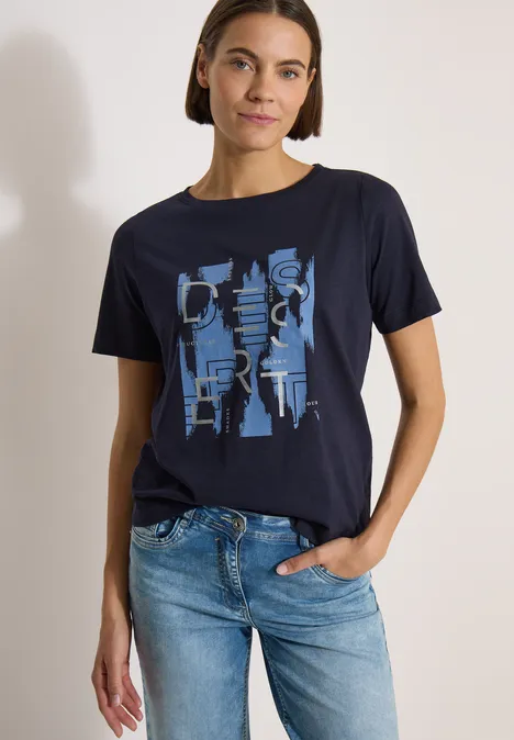 Frau trägt dunkelblaues T-Shirt mit abstraktem hellblauem Print und Jeans, eine Hand in der Hosentasche.