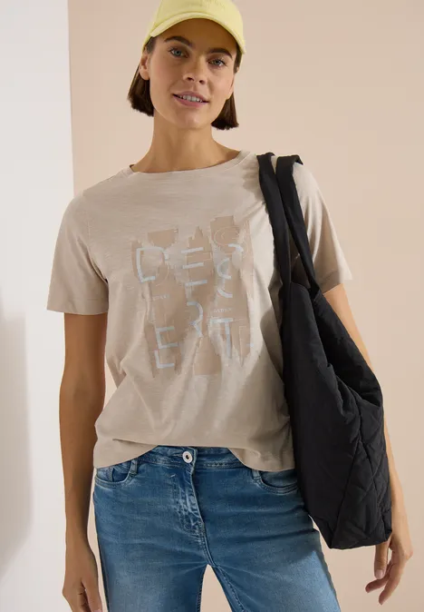 Frau trägt beige T-Shirt mit weißem Print, blaue Jeans und schwarzen, gesteppte Schulterbeutel.