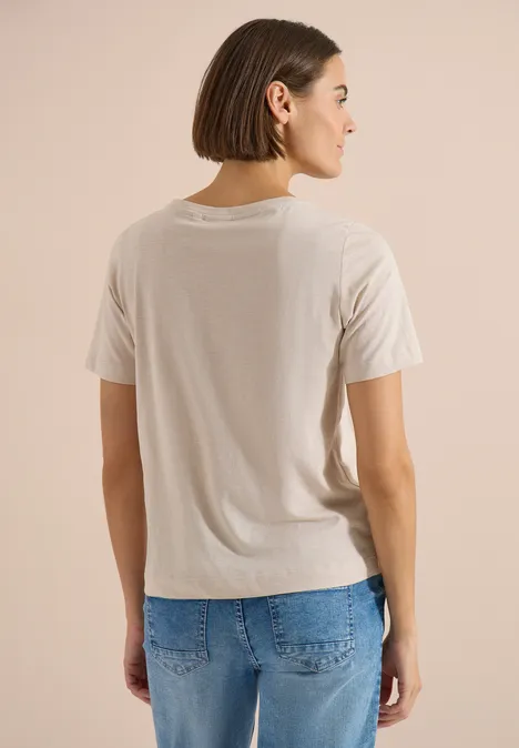 Eine Frau mit kurzen braunen Haaren trägt von hinten sichtbar ein beiges T-Shirt und eine blaue Jeans.