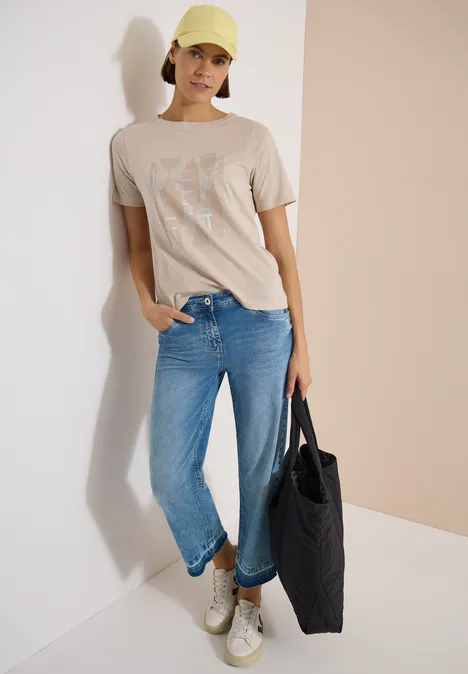 Person in beigem T-Shirt, blauen Jeans, weißen Sneakern und schwarzer Tasche.
