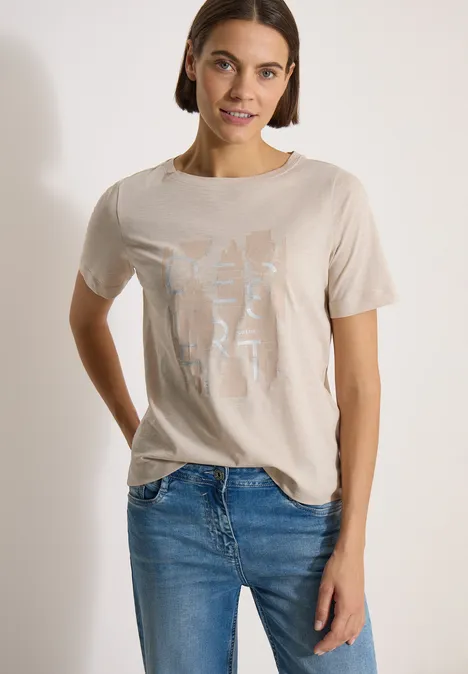 Frau in beigem T-Shirt mit hellem Aufdruck und blauen Jeans vor weißem Hintergrund.