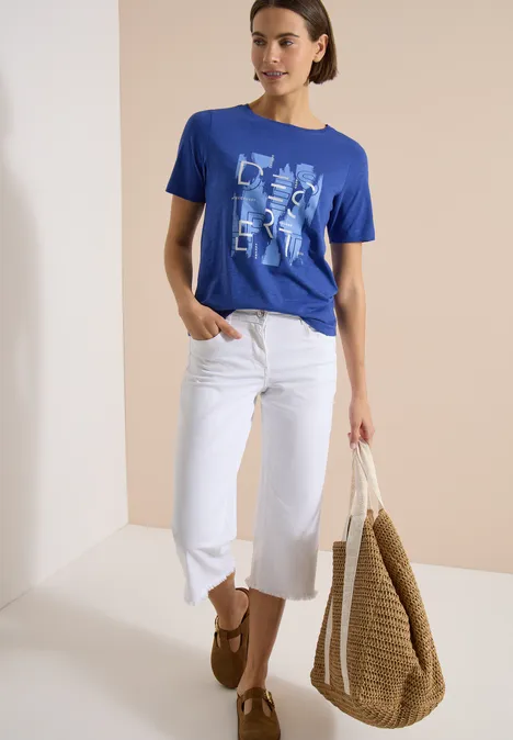 Frau trägt blaues T-Shirt mit Print, weiße Culotte-Jeans, braune Loafers und große geflochtene Korbtasche.