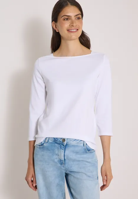 Frau in weißem Dreiviertelarm-Shirt und hellblauen Jeans lächelt seitlich vor hellem Hintergrund.