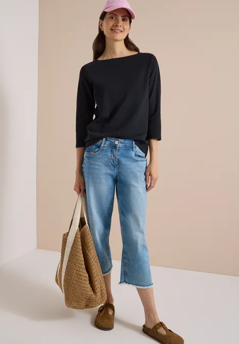 Frau mit schwarzem Shirt, hellblauen Cropped-Jeans, Strohtasche, rosa Mütze und braunen Slippern.