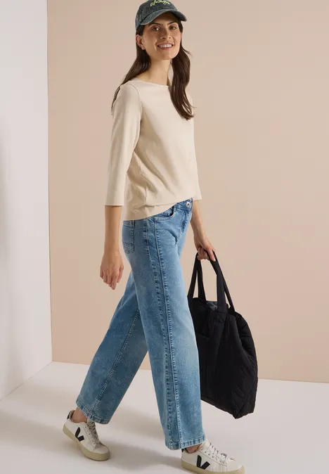 Frau mit Cap, hellbeigem Shirt, weiten Jeans und Sneakers geht lächelnd, trägt eine große schwarze Tasche.