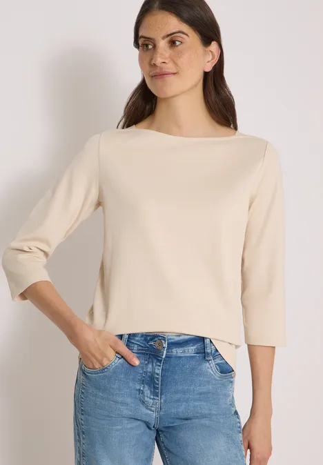 Eine Frau trägt ein hellbeiges Shirt mit 3/4-Ärmeln und blaue Jeans vor weißem Hintergrund.