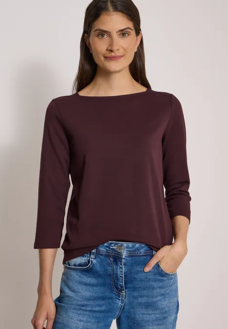 Eine lächelnde Frau in weinrotem Langarmshirt mit U-Boot-Ausschnitt und Jeans, eine Hand in der Hosentasche.