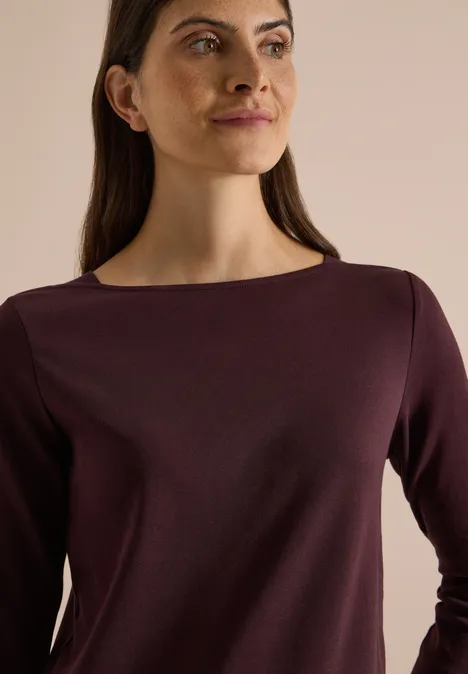 Porträt einer Frau im lila Langarmshirt, die seitlich in die Kamera blickt vor neutralem Hintergrund.