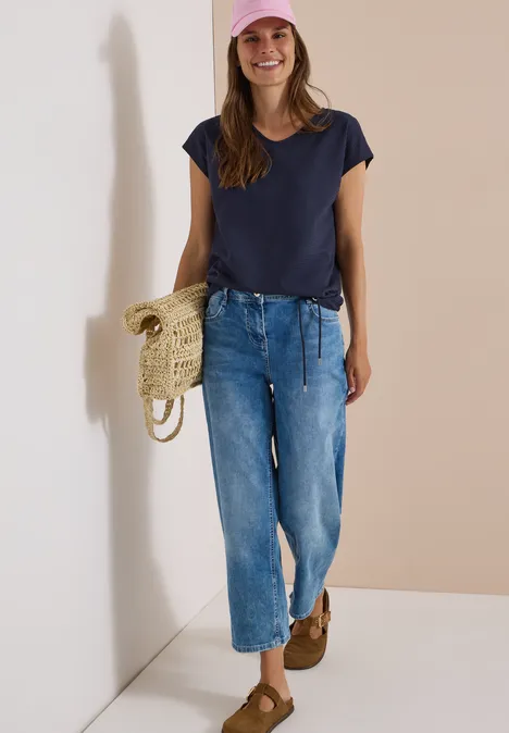 Frau in lässigem Outfit: dunkelblaues T-Shirt, weite Jeans, rosa Mütze, Strohtasche, braune Schuhe
