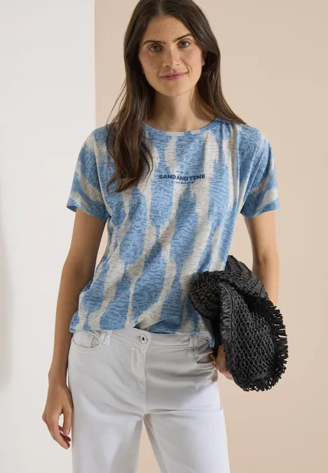 T-shirt effet délavé avec inscription urban dark blue Femme en t-shirt bleu-blanc et pantalon blanc tenant un sac noir devant un mur beige.