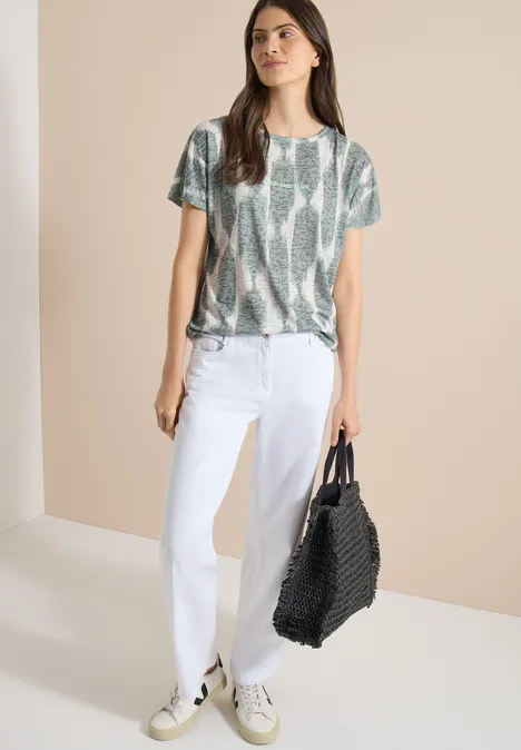 T-shirt effet délavé avec inscription deep matcha green Femme en jean blanc, blouse imprimé serpent et baskets, portant un grand sac noir tressé sur fond beige.