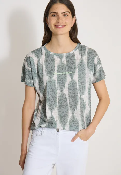 T-shirt effet délavé avec inscription deep matcha green Femme portant un t-shirt gris à motif blanc et un pantalon blanc, posant debout avec une main dans la poche.