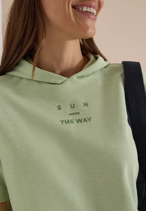 Frau mit hellem grünen Hoodie, Aufschrift 'SUN THE WAY', lächelt, schwarze Tasche über der Schulter.