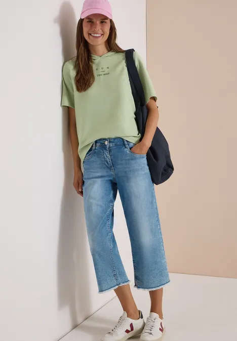 Shirt met korte mouwen, capuchon en opdruk raw matcha lime Jonge vrouw lacht tegen een muur, draagt groen T-shirt, wijd jeans, roze pet, sneakers en schoudertas.
