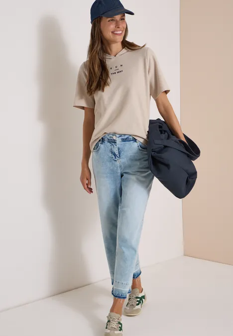 Junge Frau in beige T-Shirt, hellen Jeans und Cap, mit Jacke über dem Arm, lächelnd vor weißer Wand.