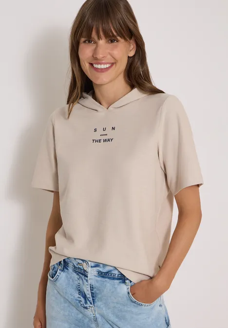 Junge Frau lächelt, trägt beiges, kurzärmliges Kapuzen-Shirt mit Aufdruck und Jeans.