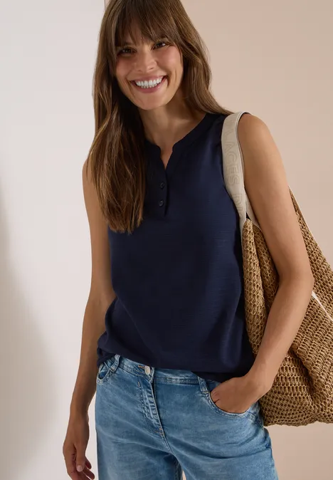 Lachende vrouw met bruin haar, blauw mouwloos top, jeans en grote gevlochten strandtas.