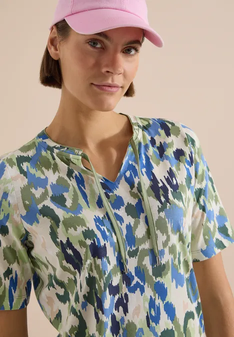 T-Shirt mit Split Neck und Print cashmere beige Person trägt kurzärmliges Kleid mit grün-blau-weißem abstraktem Muster und eine rosa Kappe.