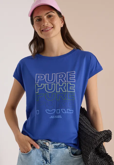 Frau in leuchtend blauem T-Shirt mit 'PURE POKE'-Print, rosa Cap, hält schwarze Strickjacke.