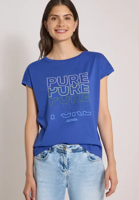 Frau in blauem T-Shirt mit Aufdruck "PURE PURE PURE" und hellen Jeans vor schlichtem Hintergrund.