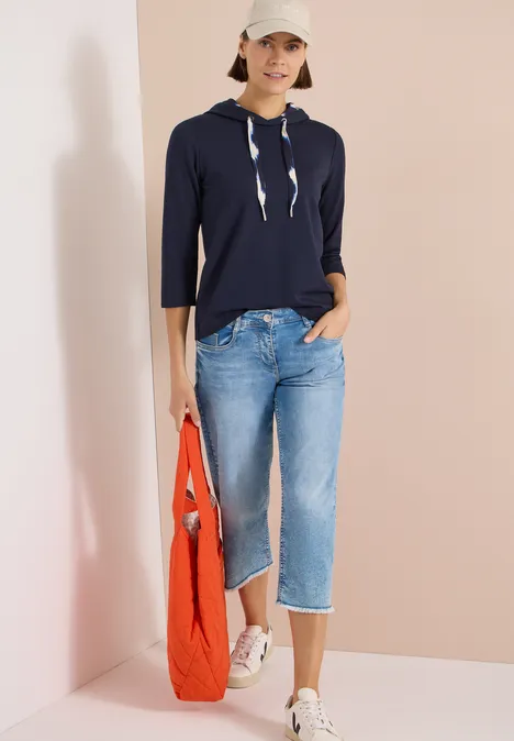 Frau trägt marineblauen Kapuzenpullover mit 3/4-Ärmeln, helle Jeans-Capris, weiße Slipper und rote Tasche.