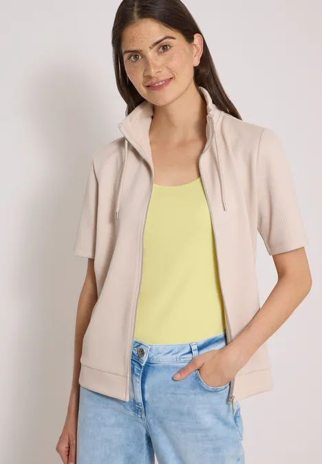 Frau trägt hellbeige Kurzarmjacke über gelbem Top und blauen Jeans, Hand in Hosentasche.