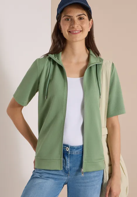 Vrouw in groene korte mouw capuchonvest, wit topje, spijkerbroek, blauw petje en tas.