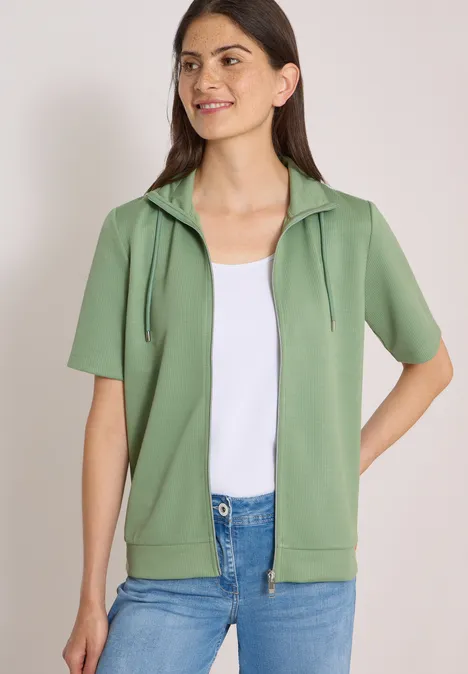 Model draagt groene korte mouw hoodie met rits, wit topje en blauwe jeans.