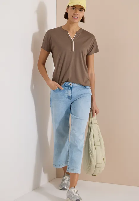 T-Shirt mit Split Neck in Unifarbe urban taupe Frau in braunem V-Ausschnitt-Shirt, hellen Jeans und Sneakern, hält beige Tasche, steht vor heller Wand.