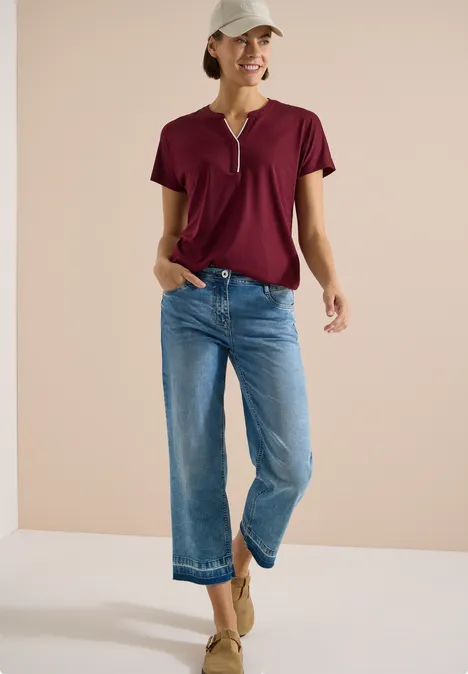 T-Shirt mit Split Neck in Unifarbe red wine Frau in rotem T-Shirt, weit geschnittenen blauen Jeans und Keilsandalen vor neutralem Hintergrund.