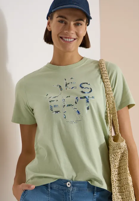 Eine Frau trägt ein hellgrünes T-Shirt mit Print, eine Netz-Tasche über der Schulter und eine dunkle Cap.