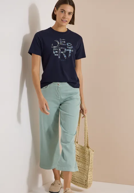 Frau steht vor neutralem Hintergrund, trägt dunkelblaues T-Shirt mit Schriftzug, mintfarbene Culotte und Basttasche.