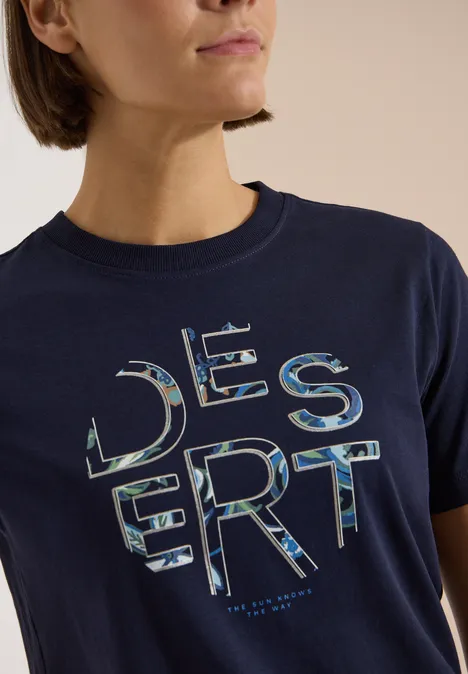 Frau trägt ein dunkelblaues T-Shirt mit 'DESERT' in camouflageähnlichen Buchstaben.