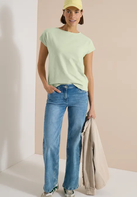 Shirt met boothals en structuur raw matcha lime Vrouw in mintgroen T-shirt, rechte blauwe jeans, draagt een beige jas, tegen een minimalistische achtergrond.
