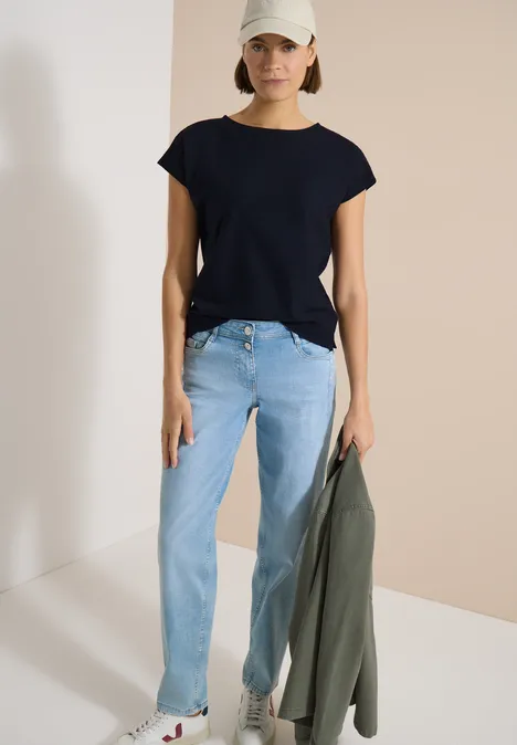 Shirt met boothals en structuur urban dark blue Vrouw in zwart T-shirt en lichte spijkerbroek houdt groene jas vast tegen lichte achtergrond.